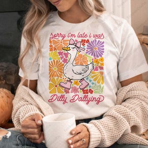 May include: White t-shirt with a colorful floral design and a cartoon goose wearing pink boots. The text "Sorry I'm late I was Dilly Dallying" is printed on the shirt.