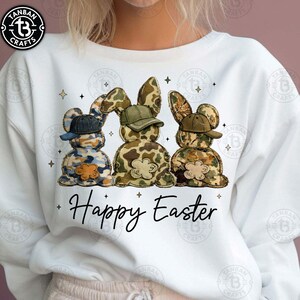Op de afbeelding: Witte sweatshirt met drie konijnenfiguren in camouflagepatronen, elk met een gekleurde pet. De tekst "Happy Easter" staat eronder in een sierlijk lettertype. De sweatshirt heeft een ronde hals. De kleuren zijn blauw, groen en bruin.