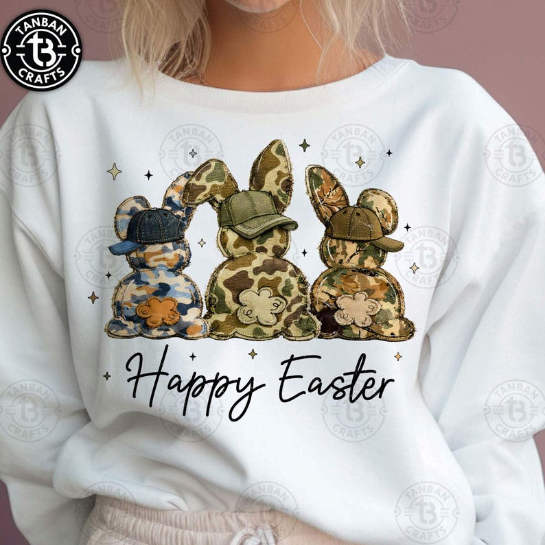 Op de afbeelding: Witte sweatshirt met drie konijnenfiguren in camouflagepatronen, elk met een gekleurde pet. De tekst "Happy Easter" staat eronder in een sierlijk lettertype. De sweatshirt heeft een ronde hals. De kleuren zijn blauw, groen en bruin.