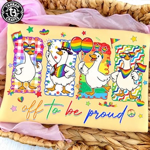 May include: A light yellow t-shirt featuring four white ducks in colorful frames. The ducks wear rainbow accessories, including hats, glasses, and scarves. The text "off to be proud" is printed in blue and rainbow colors at the bottom of the shirt.