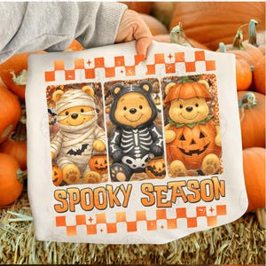 May include: A white sweatshirt featuring three rectangular panels of cartoon bears in Halloween costumes: a mummy, a skeleton, and a pumpkin. Below the panels, the text reads SPOOKY SEASON in orange letters, framed by orange checkered borders.