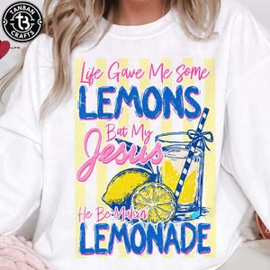 Può includere: Felpa bianca con un design grafico. Il design presenta il testo "Life Gave Me Some Lemons But My Jesus He Be-Makin' Lemonade", un bicchiere di limonata, limoni e una cannuccia.
