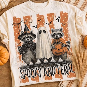 May include: A cream-colored t-shirt featuring a graphic of two raccoons and a ghost. One raccoon wears a witch hat, the other holds a jack-o'-lantern. The background has orange stripes with white dots and bats. The text reads SPOOKY AND FERAL.