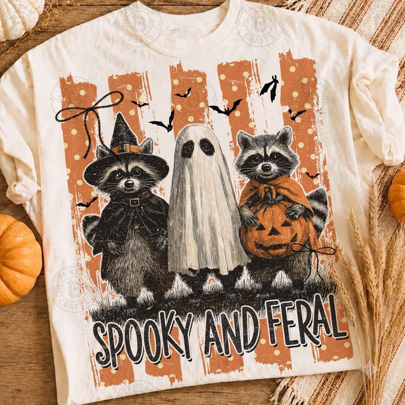 May include: A cream-colored t-shirt featuring a graphic of two raccoons and a ghost. One raccoon wears a witch hat, the other holds a jack-o'-lantern. The background has orange stripes with white dots and bats. The text reads SPOOKY AND FERAL.