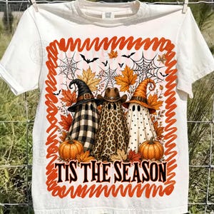 May include: A white t-shirt featuring a graphic of three ghosts wearing hats, surrounded by autumn leaves, spiderwebs, and bats. The ghosts have buffalo plaid, leopard print, and polka dot patterns. The text reads TIS THE SEASON.