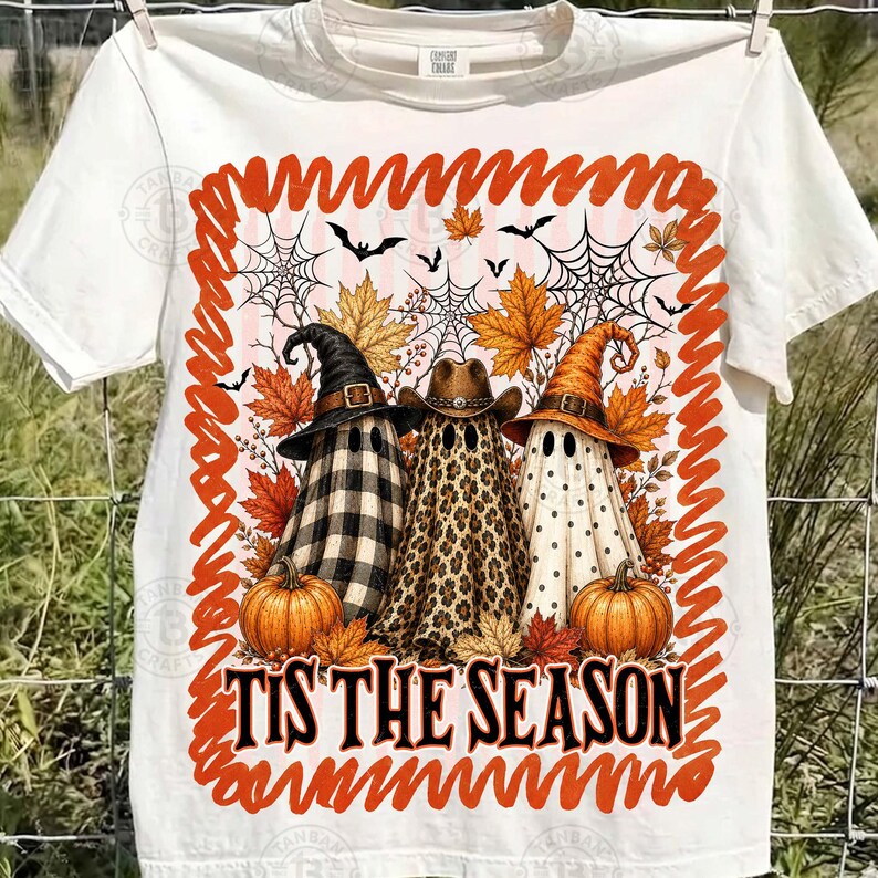 May include: A white t-shirt featuring a graphic of three ghosts wearing hats, surrounded by autumn leaves, spiderwebs, and bats. The ghosts have buffalo plaid, leopard print, and polka dot patterns. The text reads TIS THE SEASON.