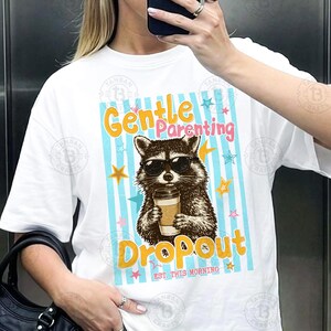 May include: White t-shirt featuring a raccoon wearing sunglasses and holding a coffee cup. The shirt has the words "Gentle Parenting Dropout" in a playful font, with stars and blue stripes. "Est. This Morning" is also printed on the shirt.