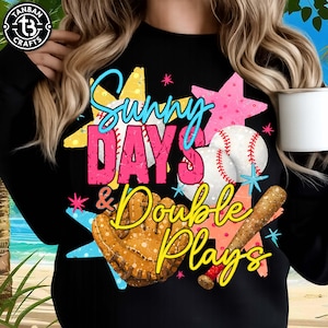 May include: Black sweatshirt with a baseball-themed graphic. The design features the words "Sunny Days & Double Plays" in a colorful, distressed font, along with a baseball, glove, and bat. The design includes yellow, pink, blue, and white accents.