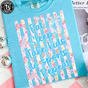 May include: A light blue t-shirt with the text "Eat Sleep Take kids to sports Repeat" in a playful font. The design includes floral stripes, stars, and sports-themed graphics. The shirt is made of a soft material.