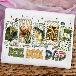 Op de afbeelding: Wit T-shirt met een visserij-thema ontwerp. Het ontwerp bevat illustraties van een tent, hengel, tacklebox, vissen en de tekst "Reel Cool Dad" met een Amerikaans vlagmotief. Het ontwerp is omlijst met groene en bruine decoratieve randen.