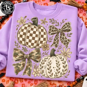 May include: A light purple sweatshirt featuring a graphic print of two pumpkins and two bows. One pumpkin has a brown and white checkered pattern, while the other has a white surface with brown leaf outlines. One bow is leopard print, the other is camouflage.