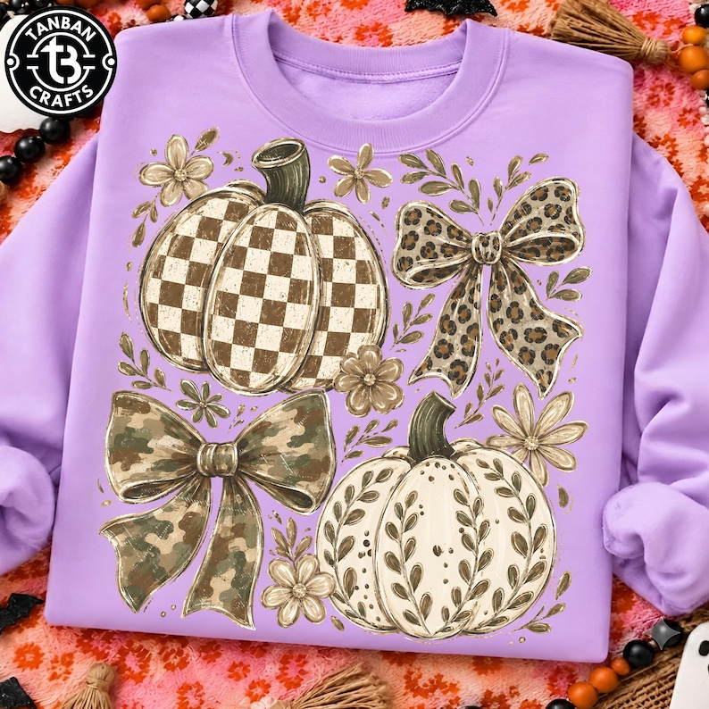 May include: A light purple sweatshirt featuring a graphic print of two pumpkins and two bows. One pumpkin has a brown and white checkered pattern, while the other has a white surface with brown leaf outlines. One bow is leopard print, the other is camouflage.