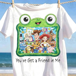 May include: A white t-shirt featuring a green frog-shaped frame graphic containing a colorful illustration of various Toy Story characters, including Woody, Buzz Lightyear, and Jessie. Below the frame, the text reads, You've Got a Friend in Me.