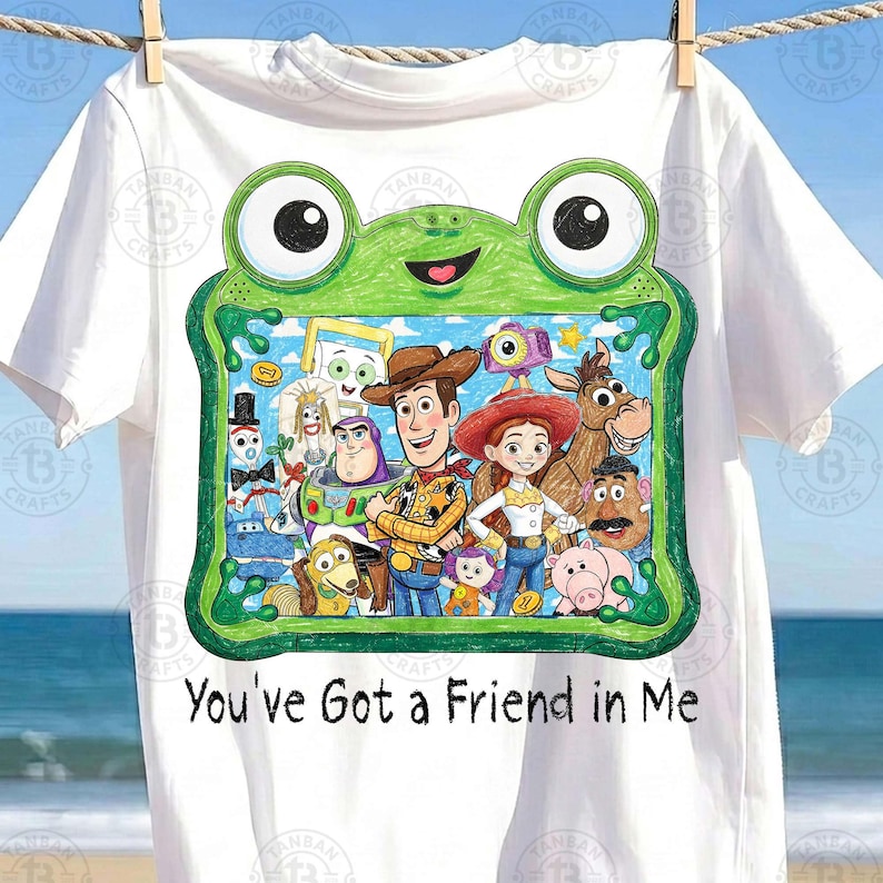May include: A white t-shirt featuring a green frog-shaped frame graphic containing a colorful illustration of various Toy Story characters, including Woody, Buzz Lightyear, and Jessie. Below the frame, the text reads, You've Got a Friend in Me.