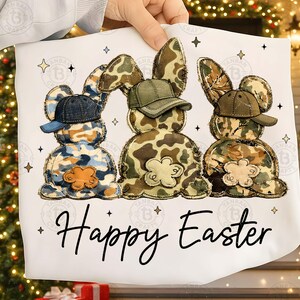 Op de afbeelding: Een wit Paaskussen met drie konijnenfiguren in camouflagepatronen, elk met een pet. De woorden "Happy Easter" staan in zwart schrift eronder. Decoratieve sterren omringen de konijnen.