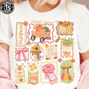 May include: A white t-shirt featuring a colorful graphic print of autumn-themed illustrations including a wagon with pumpkins, a coffee cup, a plaid scarf, sunflowers, a jar of pumpkin spread, corn, and text reading Patch picnic and Pages of Autumn.
