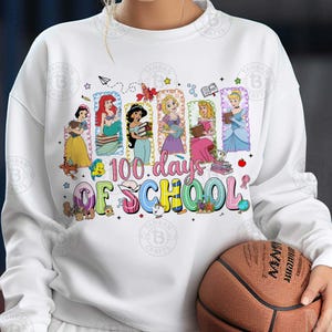 May include: White sweatshirt featuring a colorful graphic with Disney princesses and the text "100 Days of School." The princesses are depicted holding books. A basketball is held in the person's hands.
