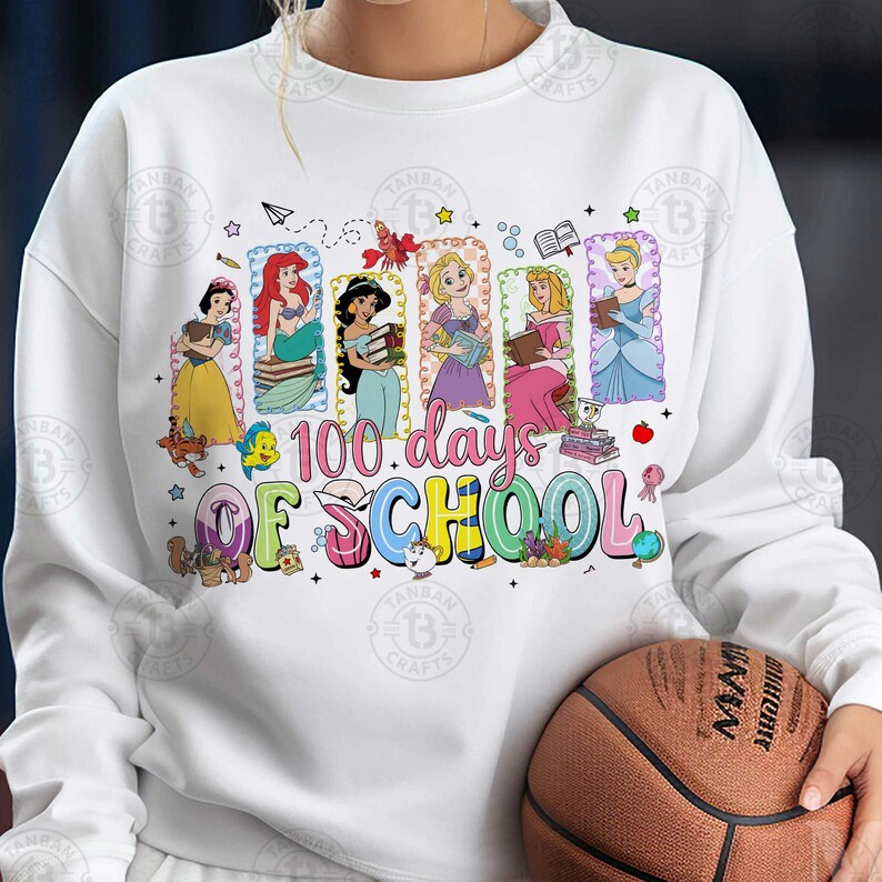 May include: White sweatshirt featuring a colorful graphic with Disney princesses and the text "100 Days of School." The princesses are depicted holding books. A basketball is held in the person's hands.