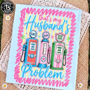 Peut inclure: T-shirt bleu clair avec un motif de trois pompes à essence vintage rouges, roses et vertes. Le texte indique "That's my Husband's Problem" en écriture bleue et rose. Le t-shirt a une bordure rose ondulée.