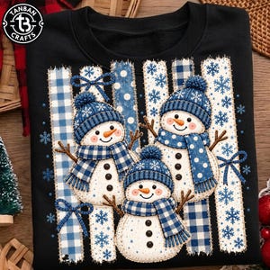 May include: A black sweatshirt featuring a graphic of three snowmen wearing blue knit hats and scarves. The snowmen are positioned against vertical stripes of blue and white plaid and polka dot patterns with blue snowflake accents.