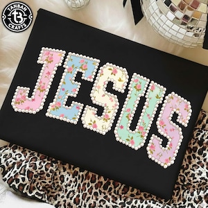 May include: Black t-shirt featuring the word "JESUS" in large, decorative letters. Each letter is filled with a floral design and edged with faux pearls. The letters are in pastel shades of pink, blue, yellow, green, and purple.