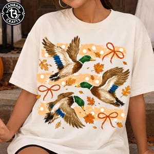 Peut inclure: Un t-shirt couleur crème arborant une impression graphique de trois canards colverts en vol. Le motif comprend des rayures orange effet coup de pinceau, des feuilles d'érable orange éparpillées et de petits nœuds orange.