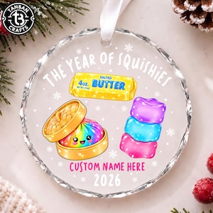 Puede incluir: Un adorno redondo de acrílico transparente con una cinta blanca, que presenta el texto THE YEAR OF SQUISHIES, CUSTOM NAME HERE y 2026. El diseño incluye una barra de mantequilla salada amarilla, una bola de masa arcoíris en una vaporera y tres squishies.