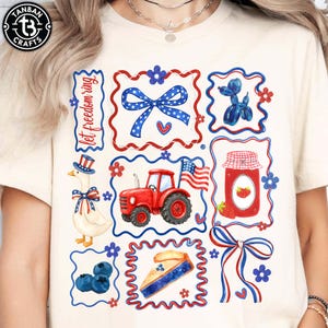 Può includere: T-shirt color crema con un design patriottico nei colori rosso, bianco e blu. Il design include un'oca, un trattore, un fiocco, una torta e un barattolo di marmellata. È presente anche il testo "let freedom ring".