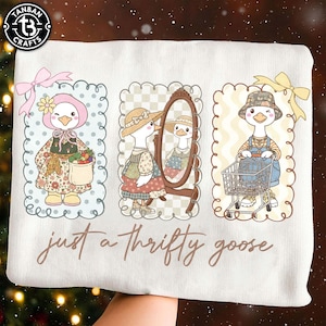 Puede incluir: Sudadera blanca con tres ilustraciones de gansos de dibujos animados. Los gansos est&aacute;n vestidos con varios atuendos, incluyendo un gorro, un vestido y un peto. El texto "just a thrifty goose" est&aacute; escrito en cursiva.