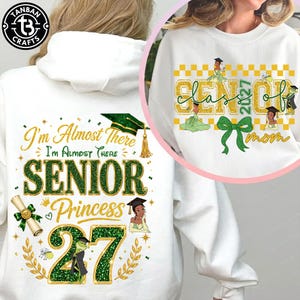 May include: A white hooded sweatshirt featuring a graphic print with the text I'm Almost There SENIOR Princess 27, a graduation cap, a diploma, and a cartoon character. A circular inset shows a white sweatshirt with SENIOR 2027 mom text.