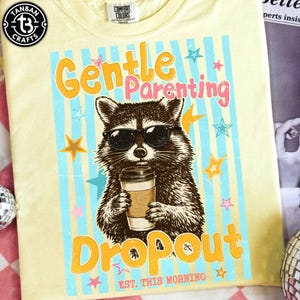 May include: A light yellow t-shirt with a graphic featuring a raccoon wearing sunglasses and holding a coffee cup. The text reads "Gentle Parenting Dropout" with stars and stripes. The shirt has a Comfort Colors tag.