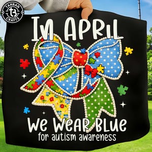 以下が含まれることがあります： 「In April we wear blue for autism awareness」のテキストが入った黒い生地。 パズルピースと水玉模様の大きなリボンが中心的なデザイン要素です。 リボンは青、赤、黄、緑の色合いです。