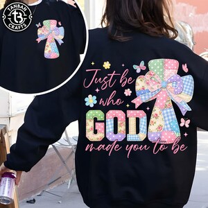 May include: Black sweatshirt featuring a colorful patchwork cross design with a bow and the text "Just be who GOD made you to be." The cross includes floral, polka dot, and gingham patterns. Butterflies and flowers are also present.