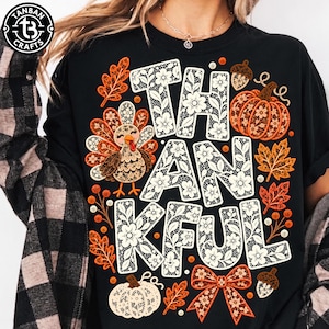 Peut inclure: Un t-shirt noir avec une impression graphique affichant le mot THANKFUL en lettres blanches à motif de dentelle. Le design comprend une dinde, des citrouilles, des feuilles d'automne, des glands et un nœud, ornés de motifs floraux en orange.