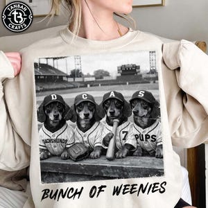 Puede incluir: Sudadera beige con un gráfico en blanco y negro de cuatro perros salchicha con uniformes y gorras de béisbol. La imagen incluye el texto "DACHSHUNDS", "7 PUPS" y "BUNCH OF WEENIES".