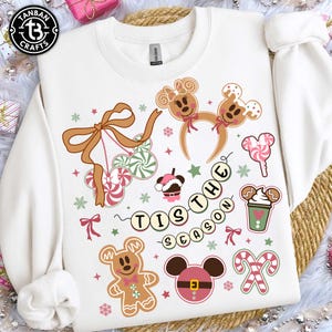 May include: A white crewneck sweatshirt featuring a festive graphic print with gingerbread characters, candy canes, peppermint swirls, and mouse-ear headbands. The text "TIS THE SEASON" is displayed in a curved banner, surrounded by small stars and snowflakes.