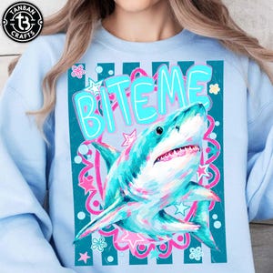 May include: Pale blue sweatshirt with a turquoise and pink shark graphic and the words "BITE ME". The shark design is detailed with turquoise and pink hues, set against a decorative background with stars and floral accents. The sweatshirt has a crew neck.