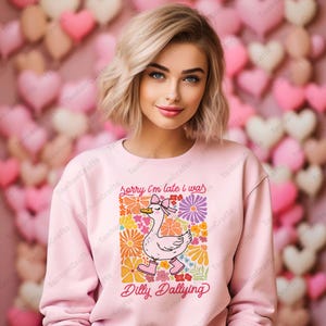 May include: A pink sweatshirt with a graphic of a cartoon duck wearing a bow and surrounded by colorful flowers. The text "Sorry I'm late I was Dilly Dallying" is printed below the duck.