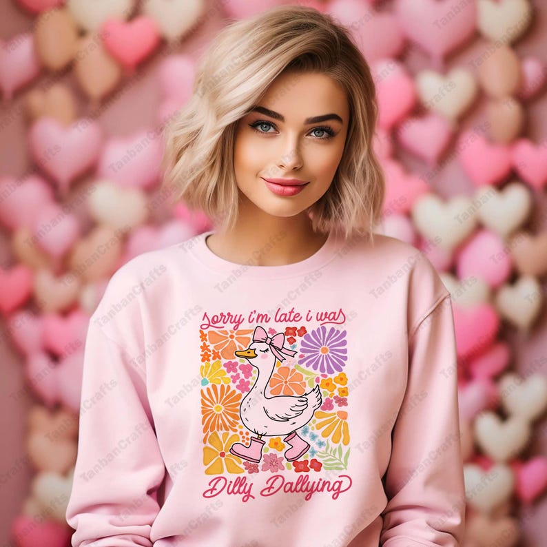 May include: A pink sweatshirt with a graphic of a cartoon duck wearing a bow and surrounded by colorful flowers. The text "Sorry I'm late I was Dilly Dallying" is printed below the duck.