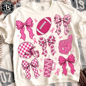 Op de afbeelding: Een off-white t-shirt met een grafische print van roze football-thema items, waaronder strikken, een football, een helm, een schuimvinger en een beker. Het ontwerp bevat lintjes voor borstkankerbewustzijn en diverse patronen.