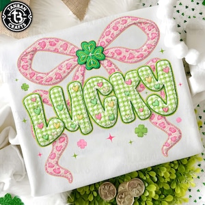 May include: White t-shirt with a large, embroidered design featuring the word "LUCKY" in green gingham letters adorned with hearts. A pink bow with a leopard print and a green shamrock complete the design. The shirt is decorated with shamrocks and stars.