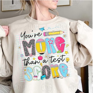 K&ouml;nnte beinhalten: Beige Sweatshirt mit dem Text "You're More Than a Test Score" in farbenfrohen, verspielten Buchstaben. Das Design beinhaltet Bleistifte, Sterne, Blumen und schulische Grafiken, perfekt f&uuml;r Lehrer oder Sch&uuml;ler.
