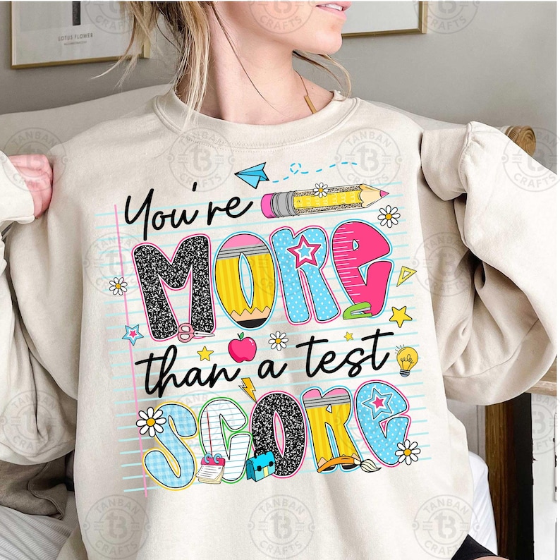 K&ouml;nnte beinhalten: Beige Sweatshirt mit dem Text "You're More Than a Test Score" in farbenfrohen, verspielten Buchstaben. Das Design beinhaltet Bleistifte, Sterne, Blumen und schulische Grafiken, perfekt f&uuml;r Lehrer oder Sch&uuml;ler.