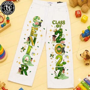May include: White denim pants featuring a colorful graphic print. The left leg displays the word SENIOR in large green letters, while the right leg shows the text CLASS OF 2027. Both legs include illustrations of a frog, a crown, and a person.