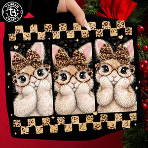 May include: Black tote bag featuring three adorable cartoon bunnies wearing glasses and leopard print bows. The bunnies have their paws up near their faces. The bag has a checkered leopard print border and the Tanban Crafts logo.