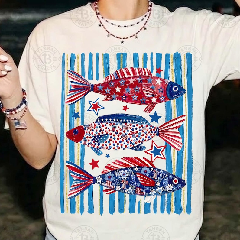 May include: White t-shirt featuring three decorative fish with patriotic designs. The fish have red, white, and blue patterns, including stars, stripes, and floral elements. Blue and white vertical stripes form a background.