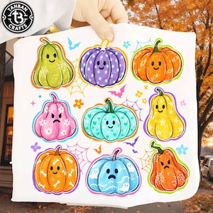May include: A white fabric square featuring a colorful graphic print of nine cartoon pumpkins with various facial expressions and patterns, including polka dots, stars, and bones, interspersed with small spiderwebs, bats, and flowers.