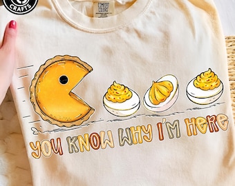 May include: A cream-colored t-shirt featuring a graphic of a yellow pie slice shaped like a video game character chasing three deviled eggs. Below the graphic, the text reads, "YOU KNOW WHY I'M HERE" in a multi-colored, rounded font.