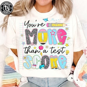 K&ouml;nnte beinhalten: Wei&szlig;es T-Shirt mit dem Text "You're MORE than a test SCORE" in farbenfrohen, verspielten Buchstaben. Das Design beinhaltet einen Bleistift, Sterne, Blumen und andere schulische Grafiken.