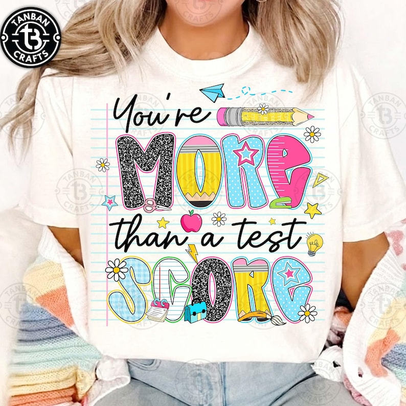 K&ouml;nnte beinhalten: Wei&szlig;es T-Shirt mit dem Text "You're MORE than a test SCORE" in farbenfrohen, verspielten Buchstaben. Das Design beinhaltet einen Bleistift, Sterne, Blumen und andere schulische Grafiken.